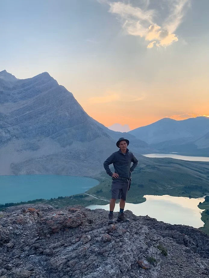 A man stands on rocky terrain in front of mountainous landscape with lakes, during sunset.