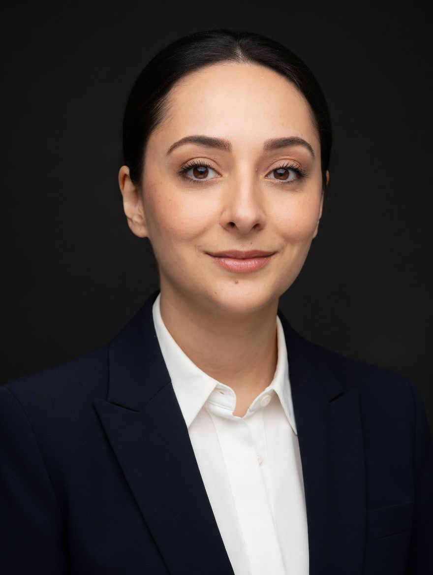 A professional young woman with dark hair, wearing a navy blazer and white shirt, smiling slightly against a dark background.