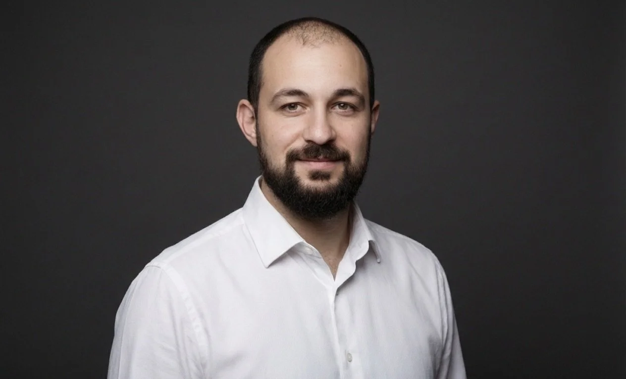 Headshot of a man with a beard and short hair wearing a white dress shirt against a dark background.