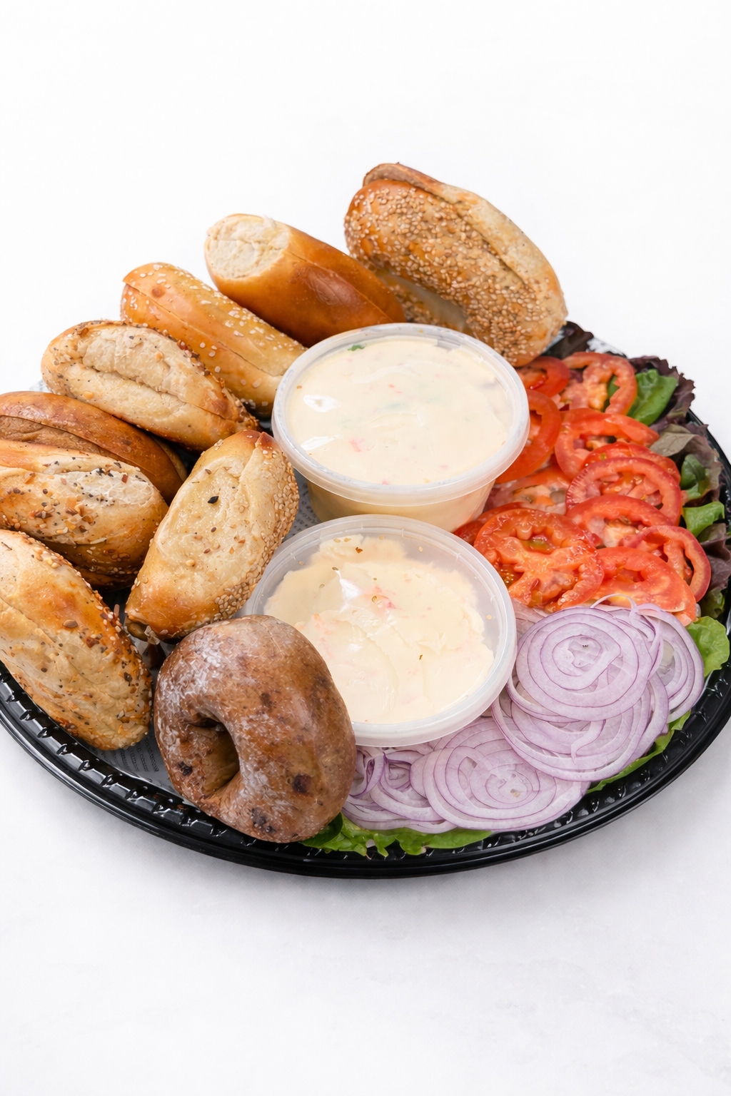 Mixed appetizer platter with assorted bagels, sliced tomatoes, sliced onions, lettuce, and two cups of creamy dipping sauces, all on a black tray.