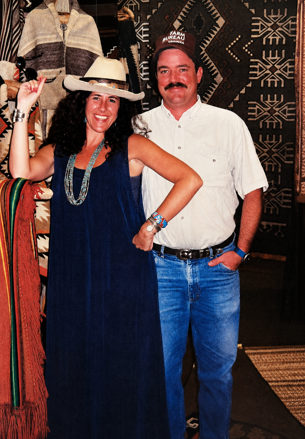 A woman wearing a beige cowboy hat, blue dress, and turquoise jewelry, and a man wearing a white shirt, blue jeans, and a red cap with the words "FARM BUREAU VIRGINIA," stand in front of a patterned textile wall. They are smiling and posing together, with the woman's arm raised and hand on her hip.