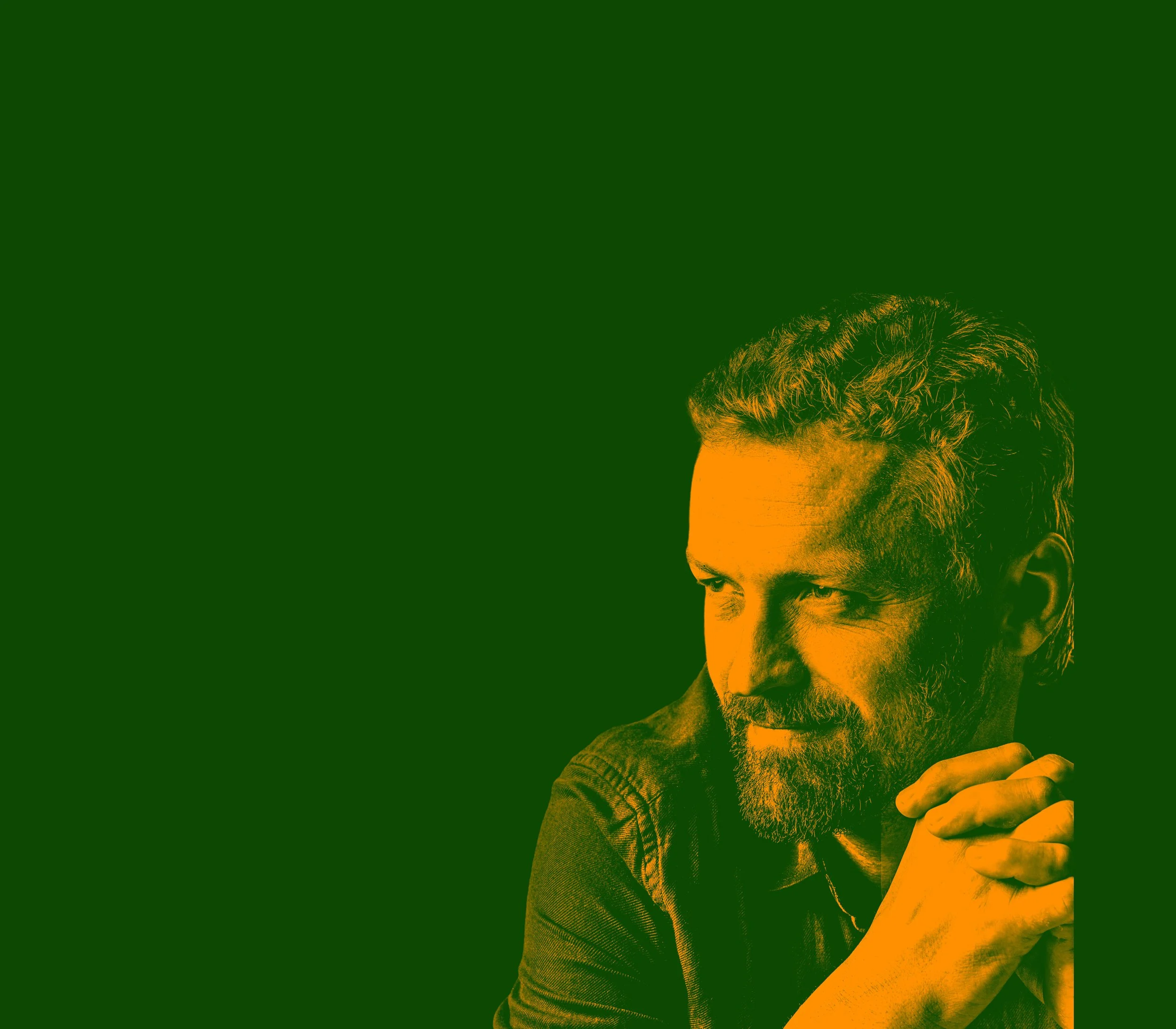 A man with a beard and curly hair, resting his chin on his folded hands, looking thoughtfully to the side against a green and orange background.