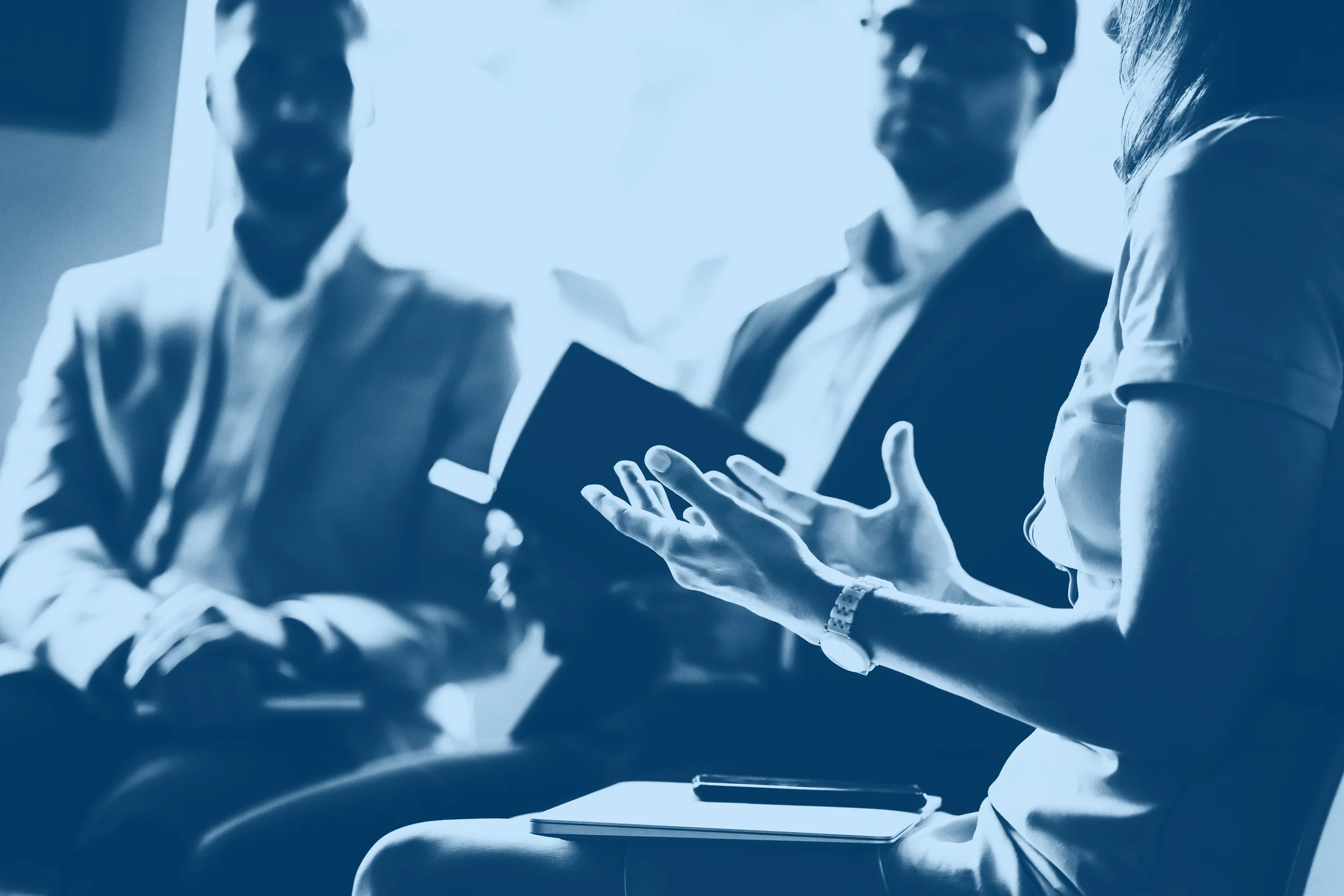 Business meeting with three people having a discussion, one woman is speaking and gesturing with her hands, the others are listening attentively, in a professional setting.
