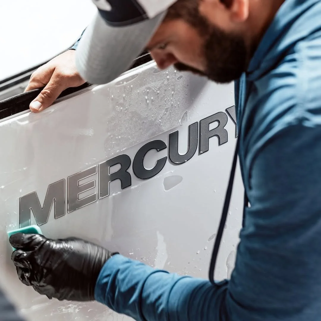A man in a blue hoodie and black gloves is cleaning the side of a white boat or watercraft with the word 'Mercury' on it, using a small sponge.