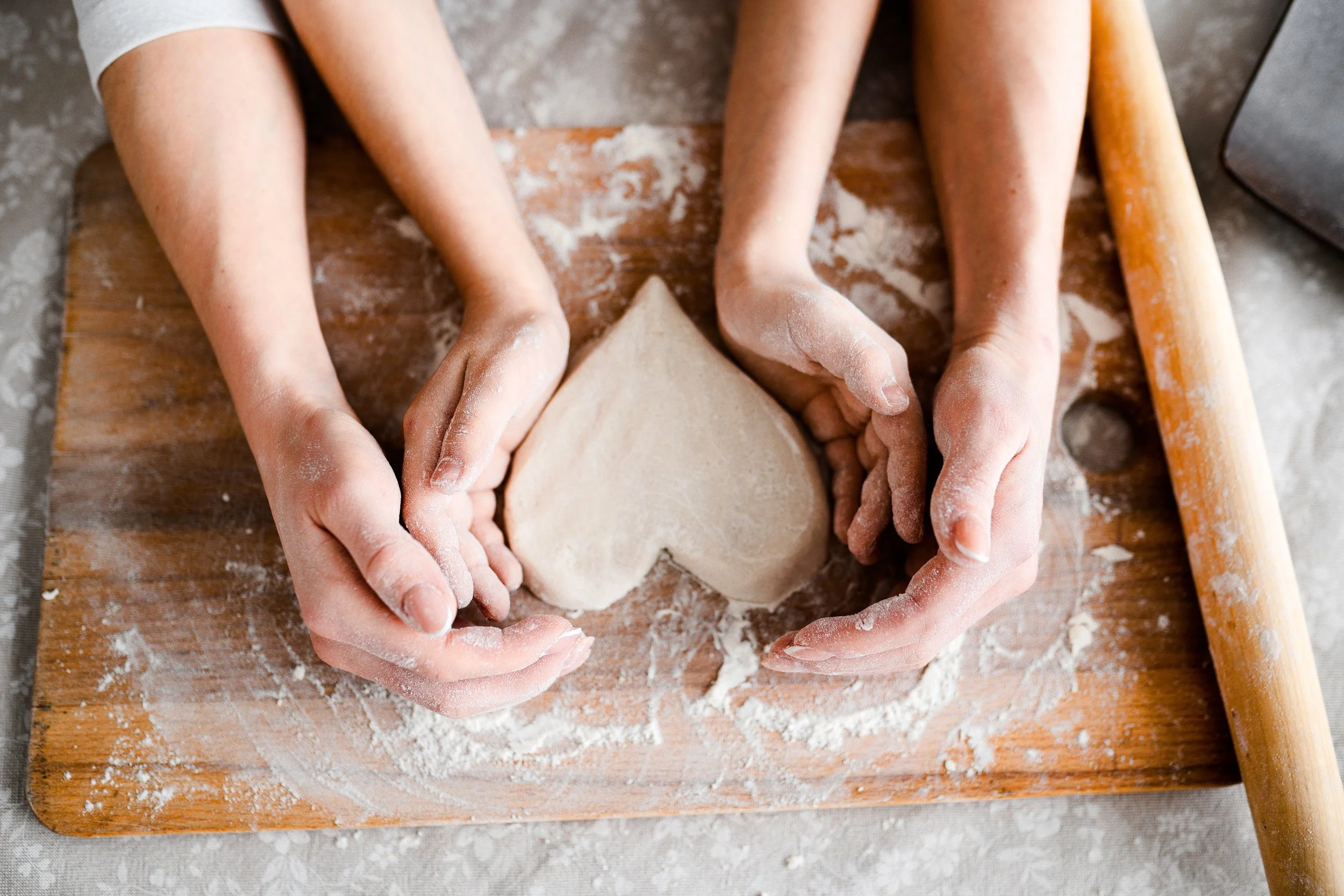 Looking down at a marble surface that has a cutting board covered with flour on top and a rolling pin sitting to the right. A pair of adult hands are cupping a pair of child hands, in the middle sits an unbaked, heart-shaped scone.