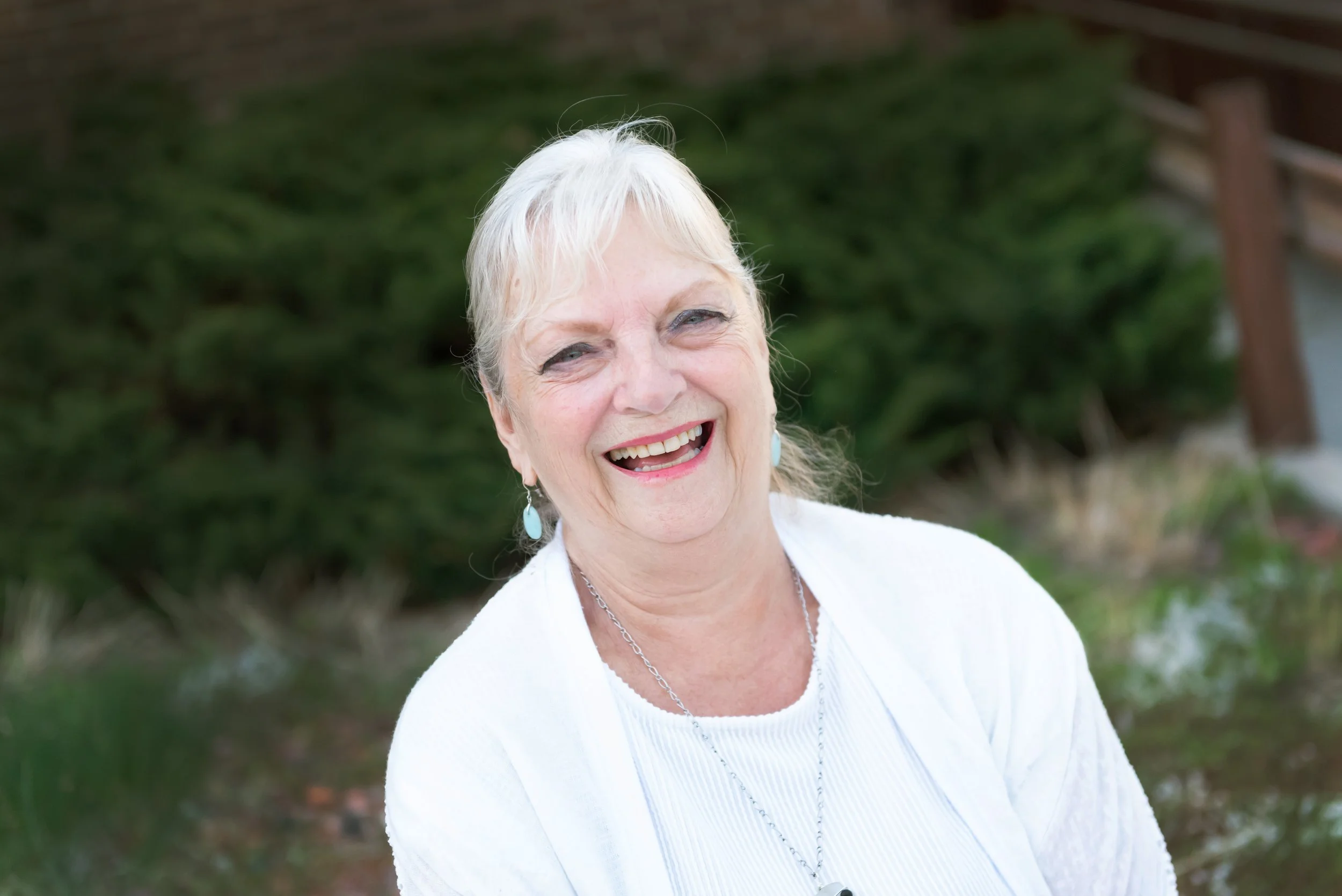A smiling senior woman with short gray hair, wearing earrings and a white top, outdoors with greenery in the background.