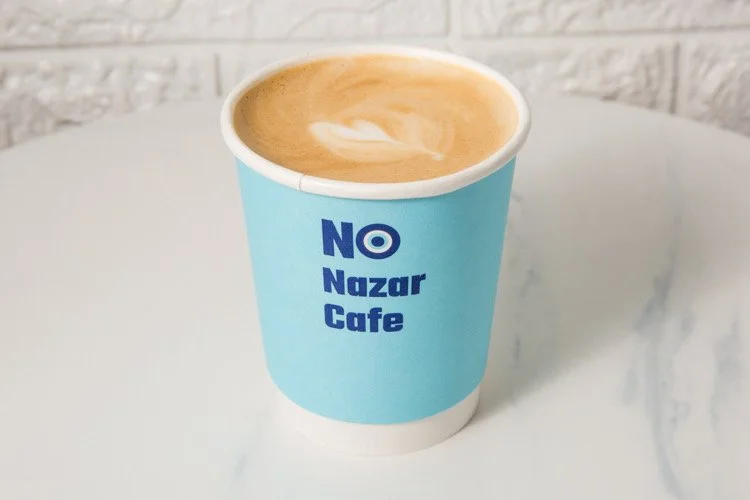 A blue paper coffee cup with the words "NO Nazar Cafe" written on it, filled with a latte or cappuccino with foam on top, placed on a white table.