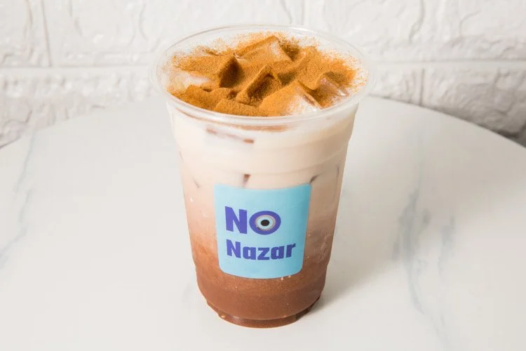 A plastic cup of iced coffee topped with cinnamon and a label reading 'NO Nazar' placed on a white marble surface.