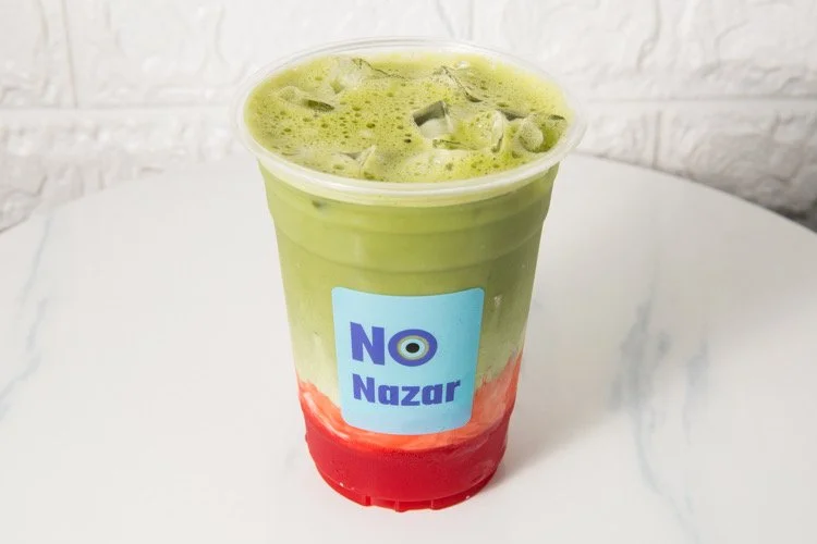 Cold beverage with green, white, and red layers in a clear plastic cup, topped with ice, placed on a white surface.