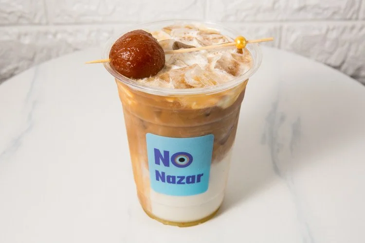 A plastic cup with a blue label reading 'NO Nazar'. The drink inside appears to be iced coffee or tea with milk, topped with ice and a cherry on a skewer.