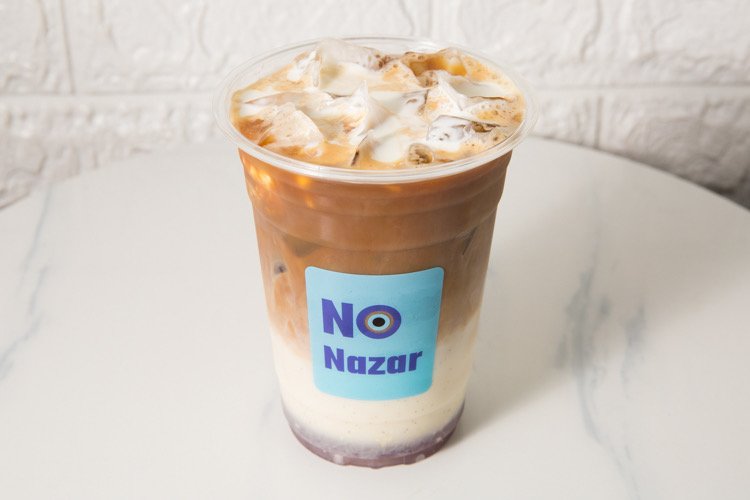 Iced coffee or latte with milk and coffee layers in a clear plastic cup with a NO Nazar logo, on a white marble surface.