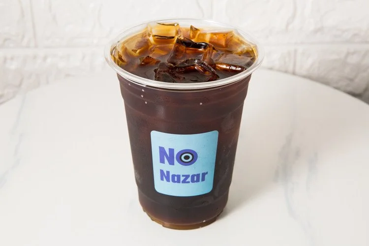 A clear plastic cup filled with iced coffee topped with ice cubes, displaying a blue and white label that says 'No Nazar'.