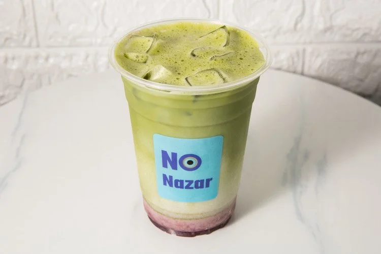 Ice green tea latte with a pink base in a clear plastic cup with a blue label reading 'NO Nazar', placed on a white surface with a textured white wall in the background.