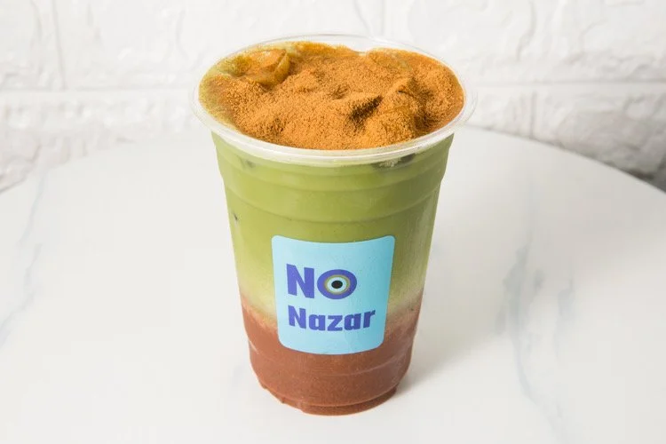 A plastic cup with a layered drink featuring green, white, and brown sections, topped with a brown powder, and labeled with a blue sticker that says 'NO Nazar'.