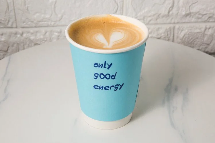 A blue paper coffee cup with the words "only good energy" in blue font, filled with a latte featuring heart-shaped foam art, placed on a white marble table.