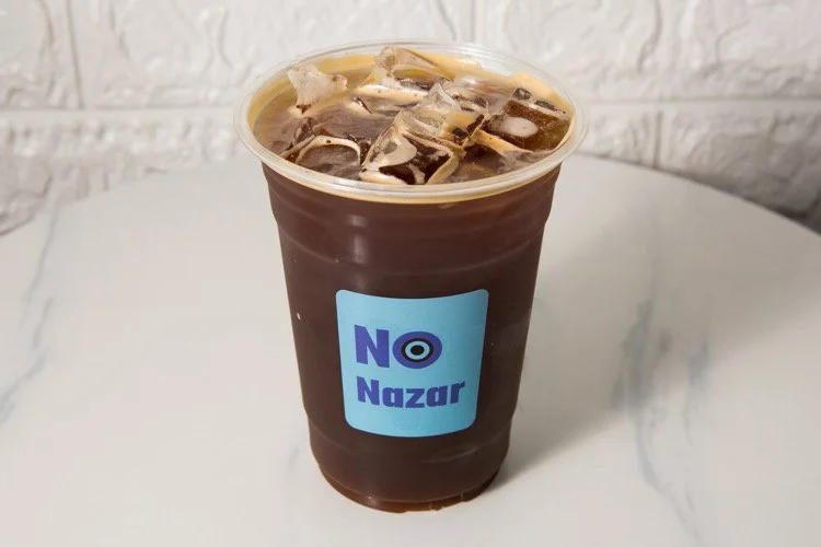 Plastic cup filled with iced coffee, displaying the logo of 'No Nazar' on the front, placed on a white marble surface with a white textured wall in the background.