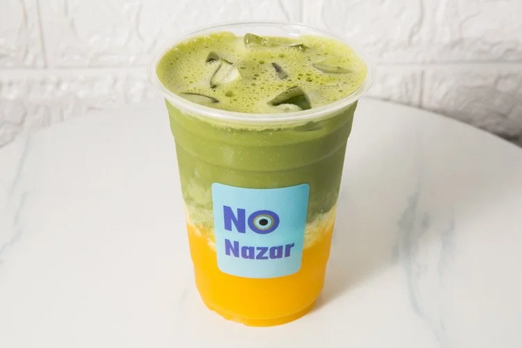 Two-layer cold beverage in a plastic cup with ice, a green top layer and a yellow bottom layer, and a label that says "NO Nazza".