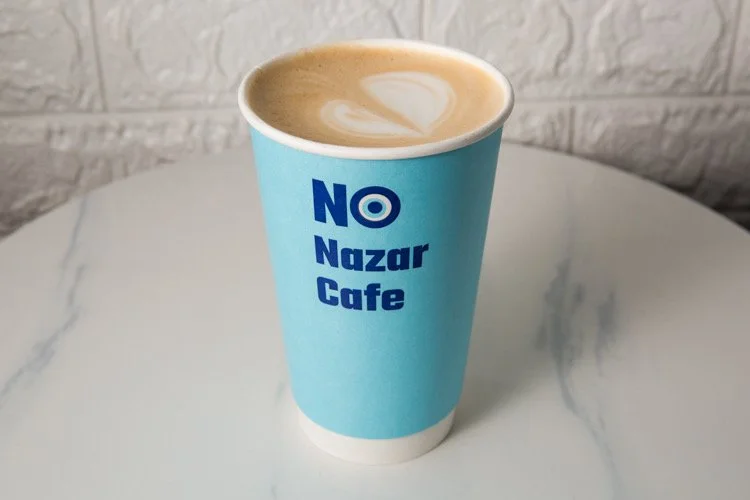 A blue paper coffee cup with the text "NO Nazar Cafe" on it, filled with a latte featuring a latte art heart on top, placed on a white marble table against a white brick wall background.