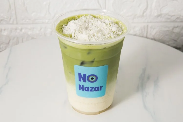 Layered iced drink with green tea, white cream, and shredded topping in a clear plastic cup labeled 'NO Nazar' on a white marble surface.