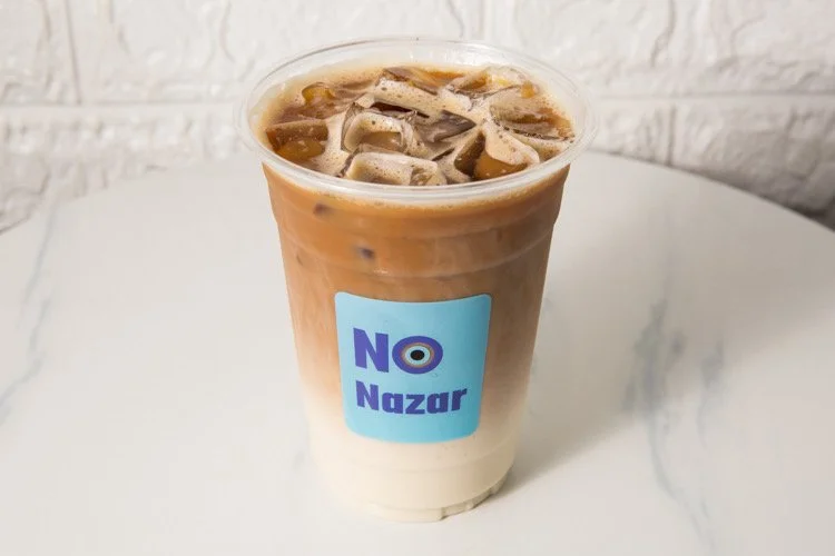 Iced coffee in a clear plastic cup with ice, featuring a blue and purple label that says "NO Nazar". The coffee appears to have layers of milk or cream at the bottom.