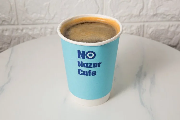 A disposable blue coffee cup with the words 'NO Nazar Cafe' printed on it, filled with coffee, placed on a white marble table.
