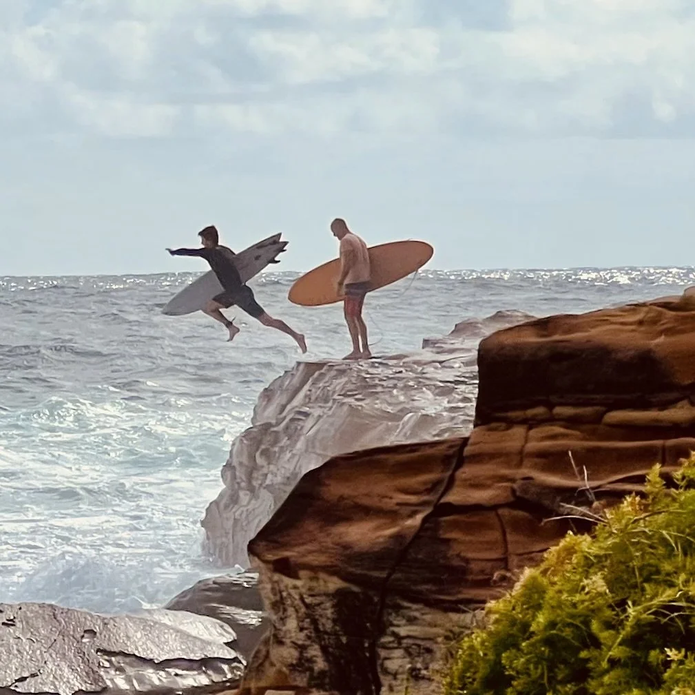Two surfers on rocks by the ocean, one jumping with a surfboard and the other standing with a surfboard.