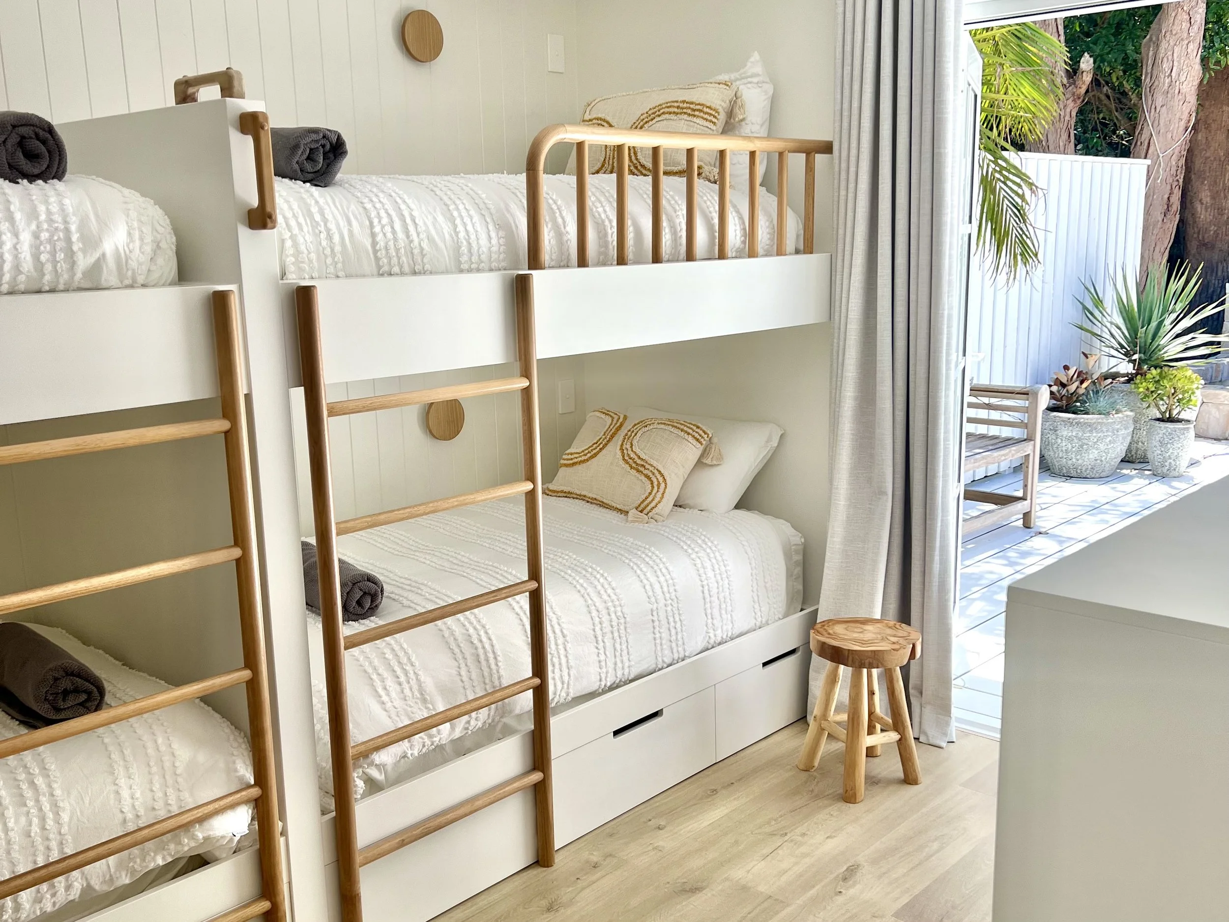 A bedroom with white bunk beds featuring wooden ladders and guard rails, white bedding with embroidered details, a small wooden stool, and an outdoor patio with potted plants and seating visible through open curtains.