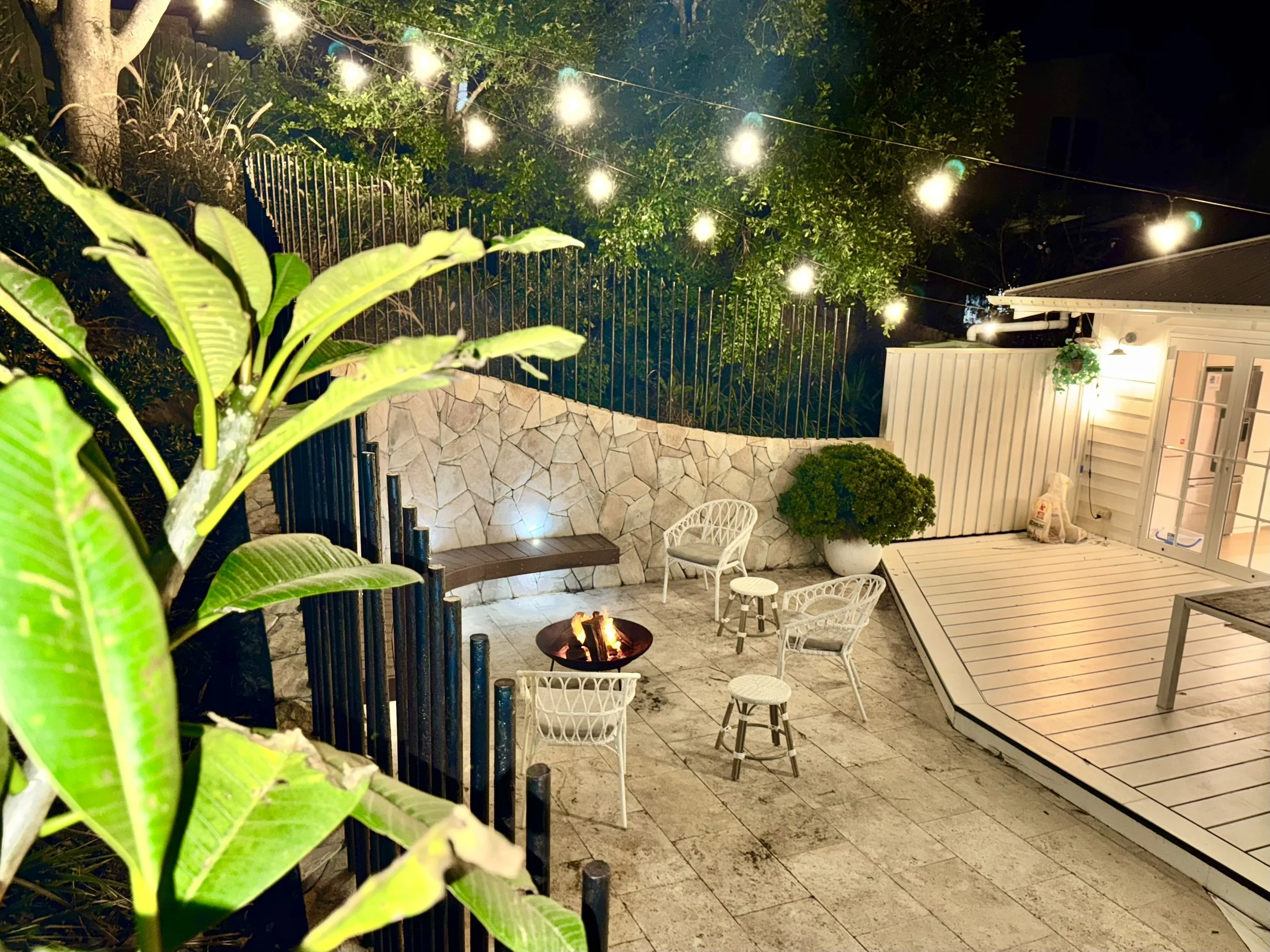 A cozy outdoor patio at night with string lights, a fire pit, white chairs, potted plants, a stone wall, a deck, and a house with a door and window.
