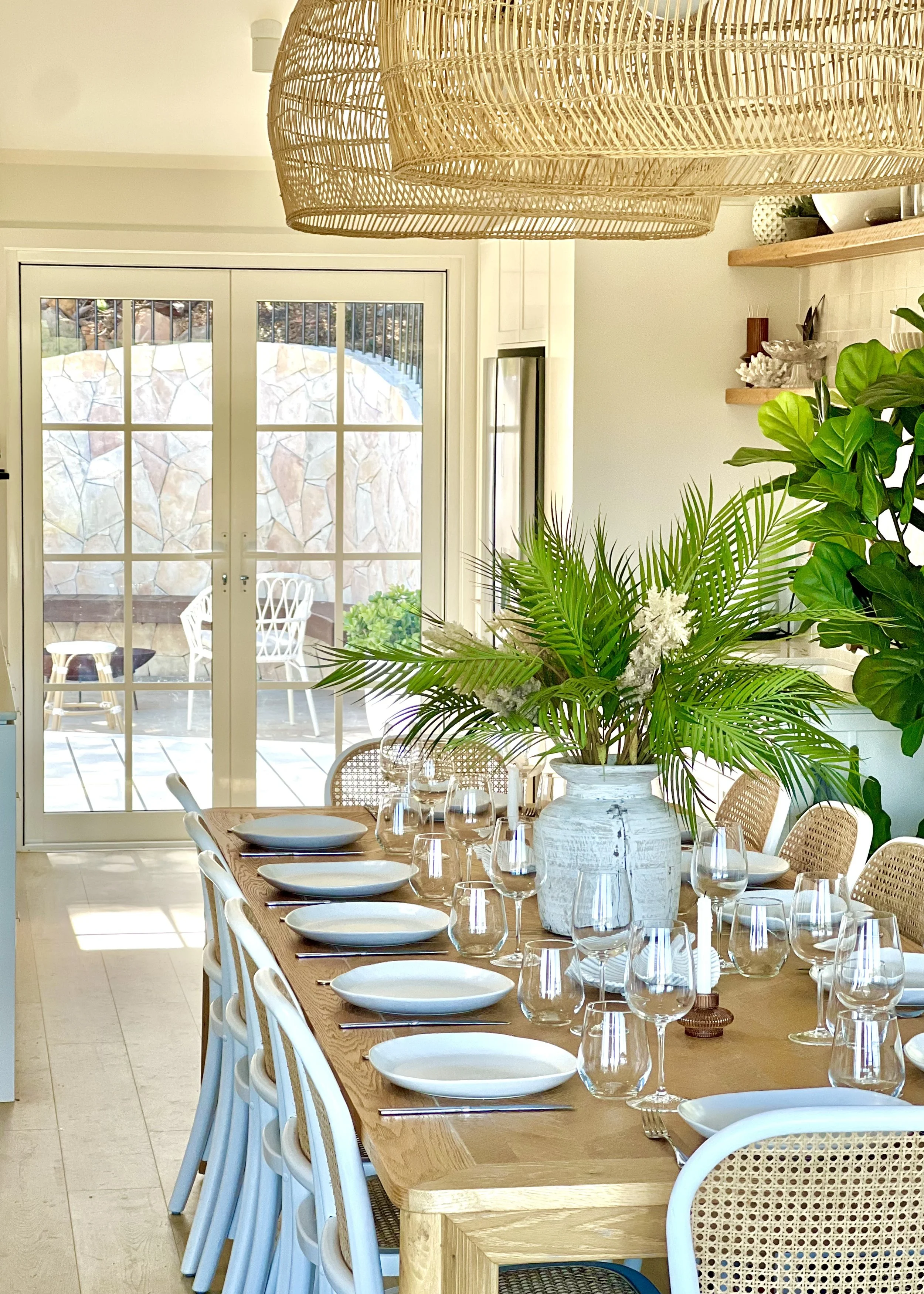 Dining room with a wooden table set for a meal, with plates, wine glasses, and cutlery. The table has a large white vase with green tropical plants. Sunlight streams in through glass doors that lead to a patio with outdoor furniture, and there are be