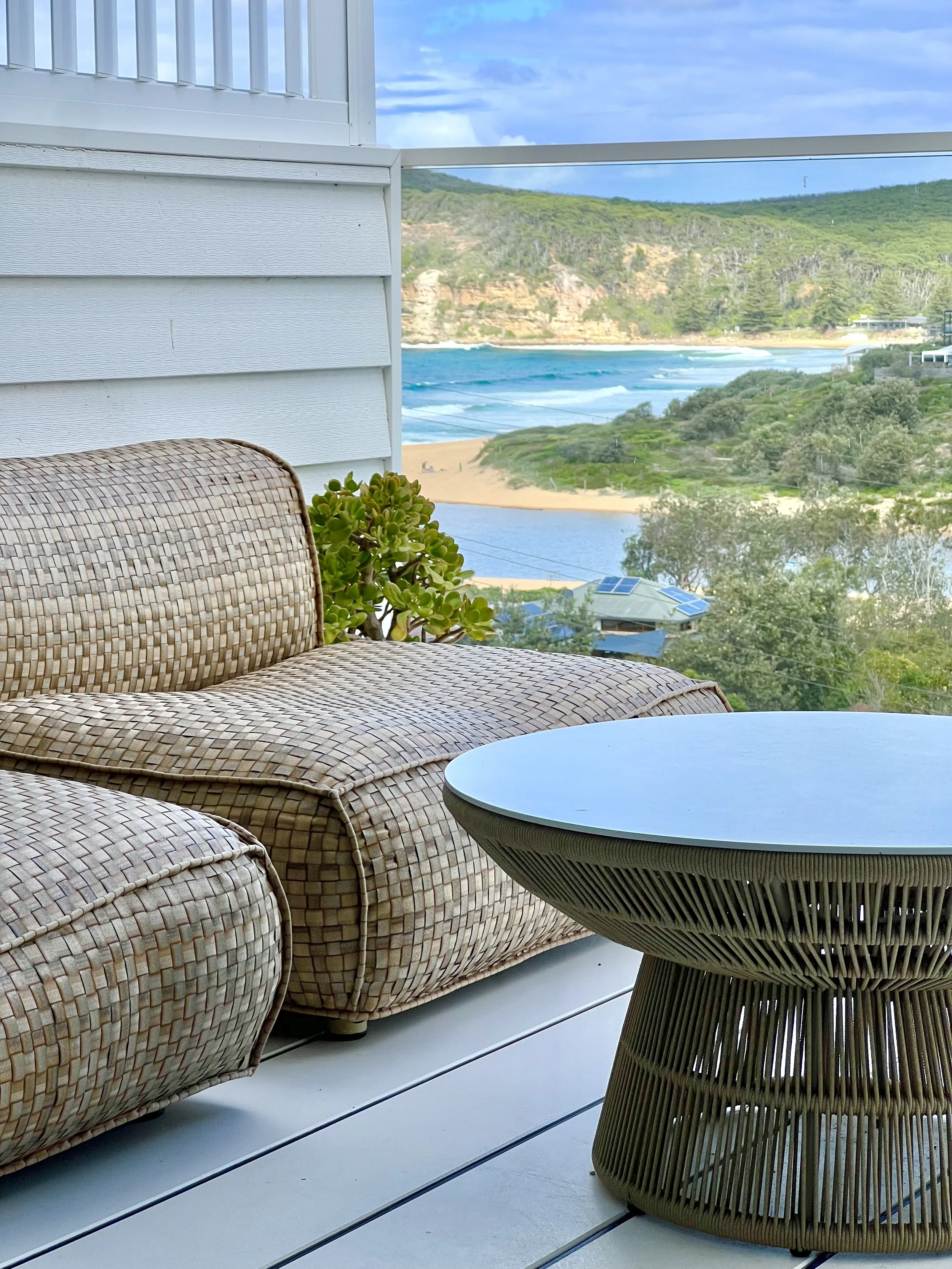 Outdoor balcony with rattan sofa, small round table, and a view of the beach, ocean, and greenery.