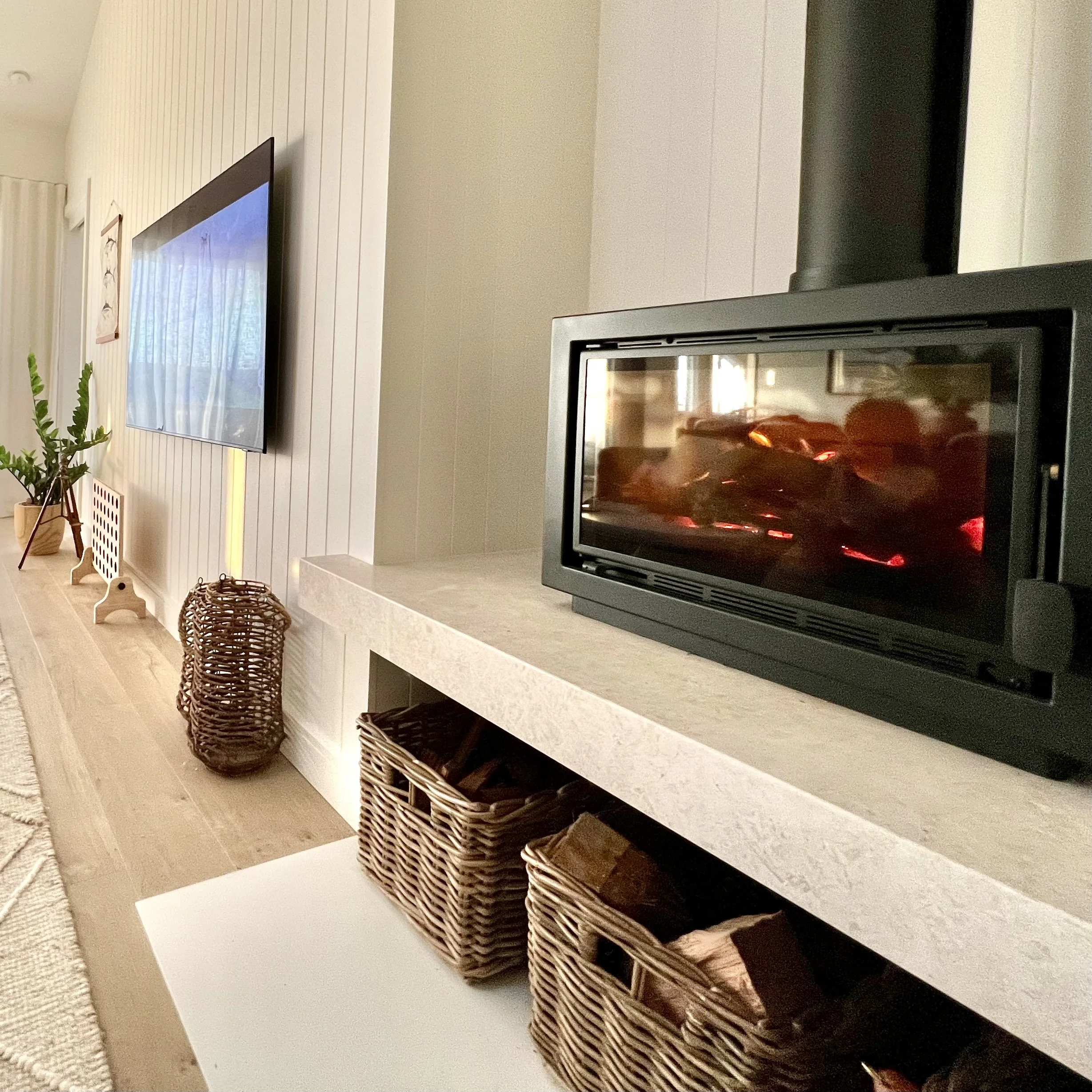 Living room with a wall-mounted flat-screen TV, a white stone fireplace with a wood stove, and woven baskets on the floor near the fireplace. There is a green plant in a pot and a window with curtains in the background.