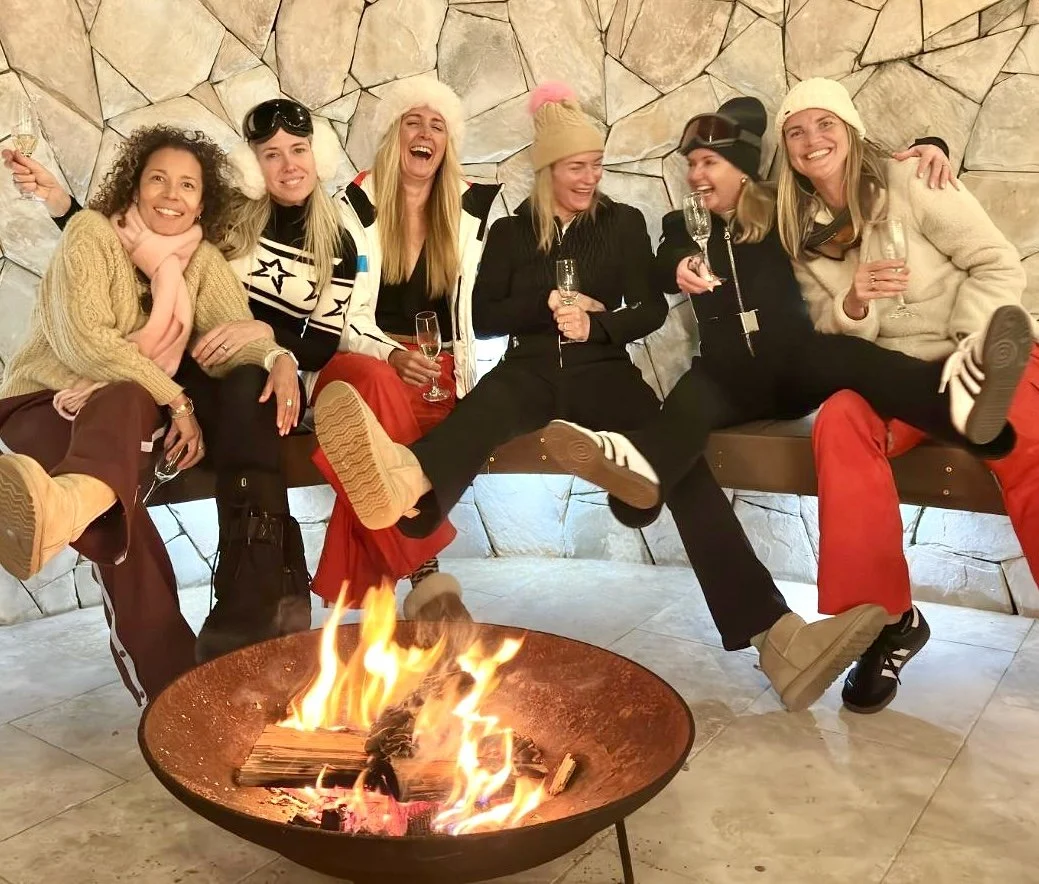 Girls weekend away on the central coast in Copacabana. Group of six women sitting and laughing around a fire pit, holding glasses of champagne, dressed warmly with winter hats and sweaters.
