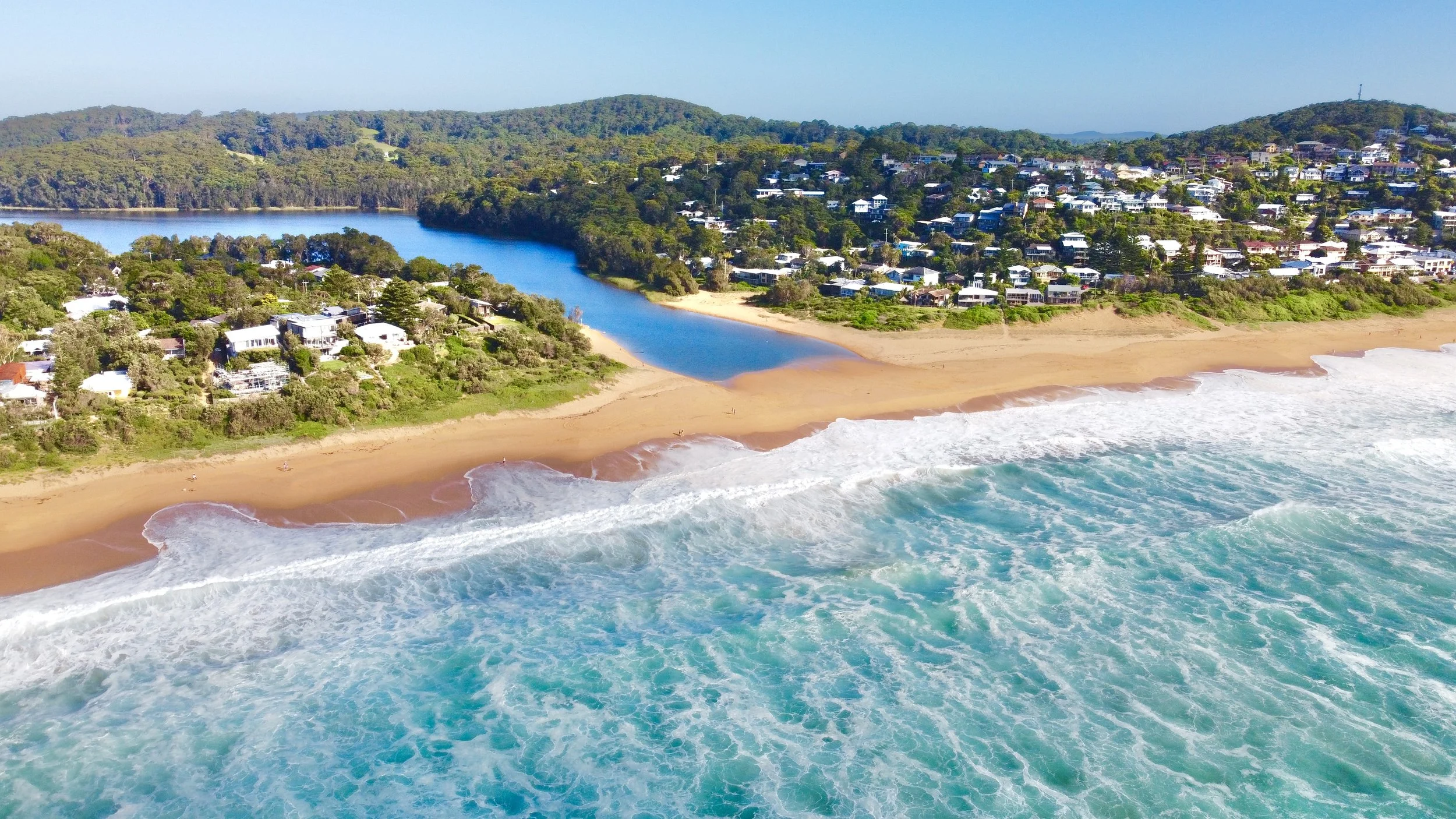 Aerial view of a coastline with sandy beach, blue ocean waves, residential houses, a river flowing into the ocean, and forested hills in the background.