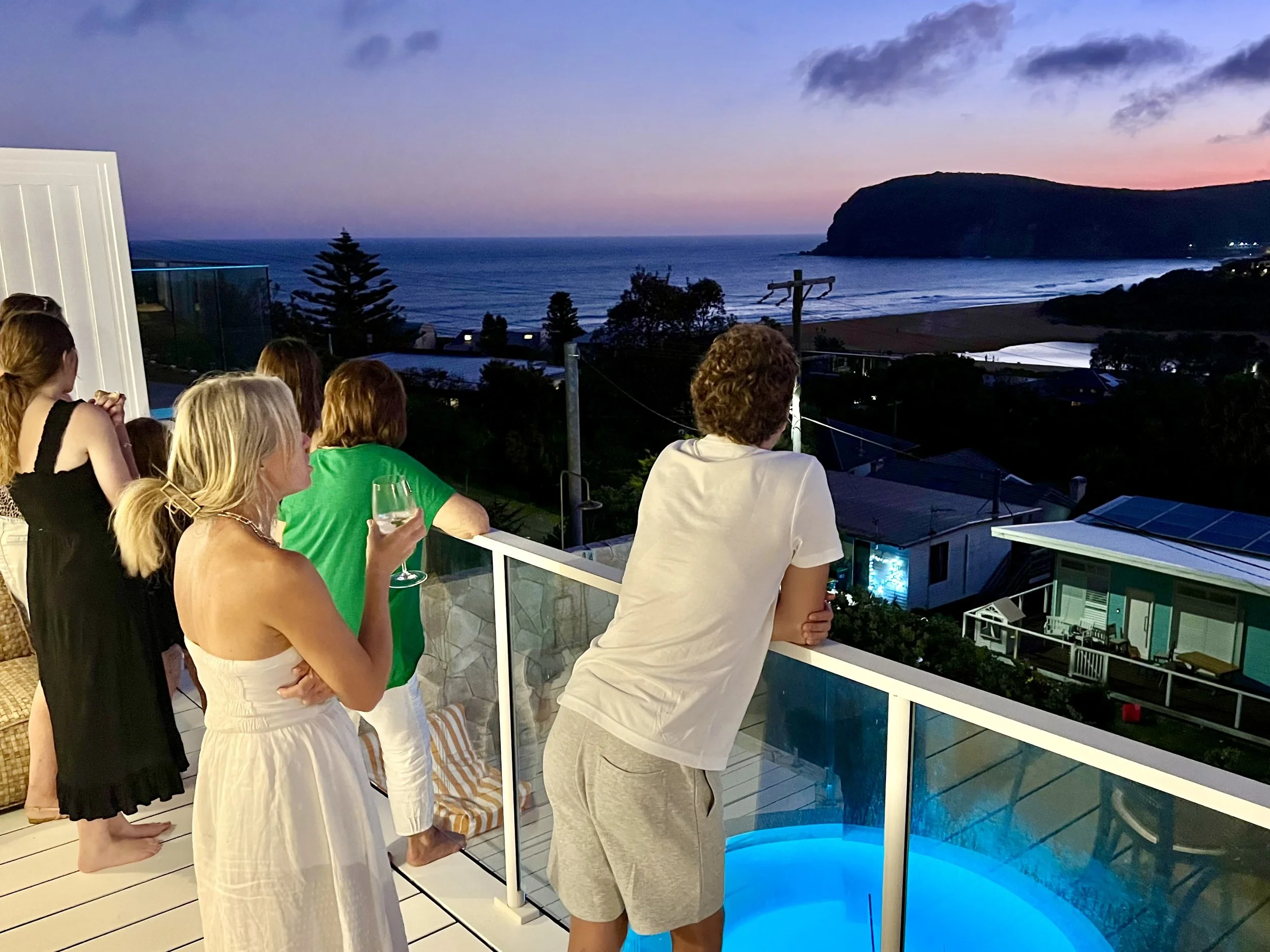 Group of people on a balcony overlooking a beach at sunset, some holding drinks and gazing at the view.