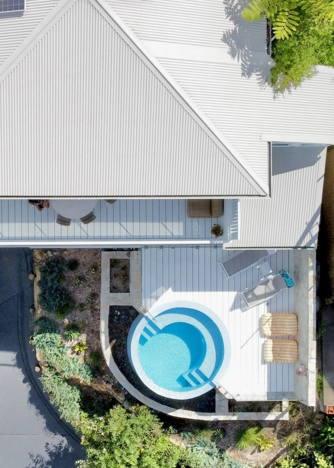 Aerial view of a backyard with a round swimming pool, white deck, lounge chairs, and surrounding landscaping with plants and trees.