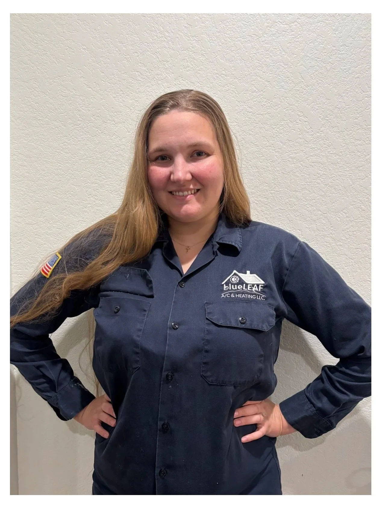 A woman with long blond hair wearing a dark blue work uniform with a patch on the sleeve and a logo on the chest that reads 'blueLEAF A/C & HEATING LLC' stands with her hands on her hips, smiling, in front of a plain wall.