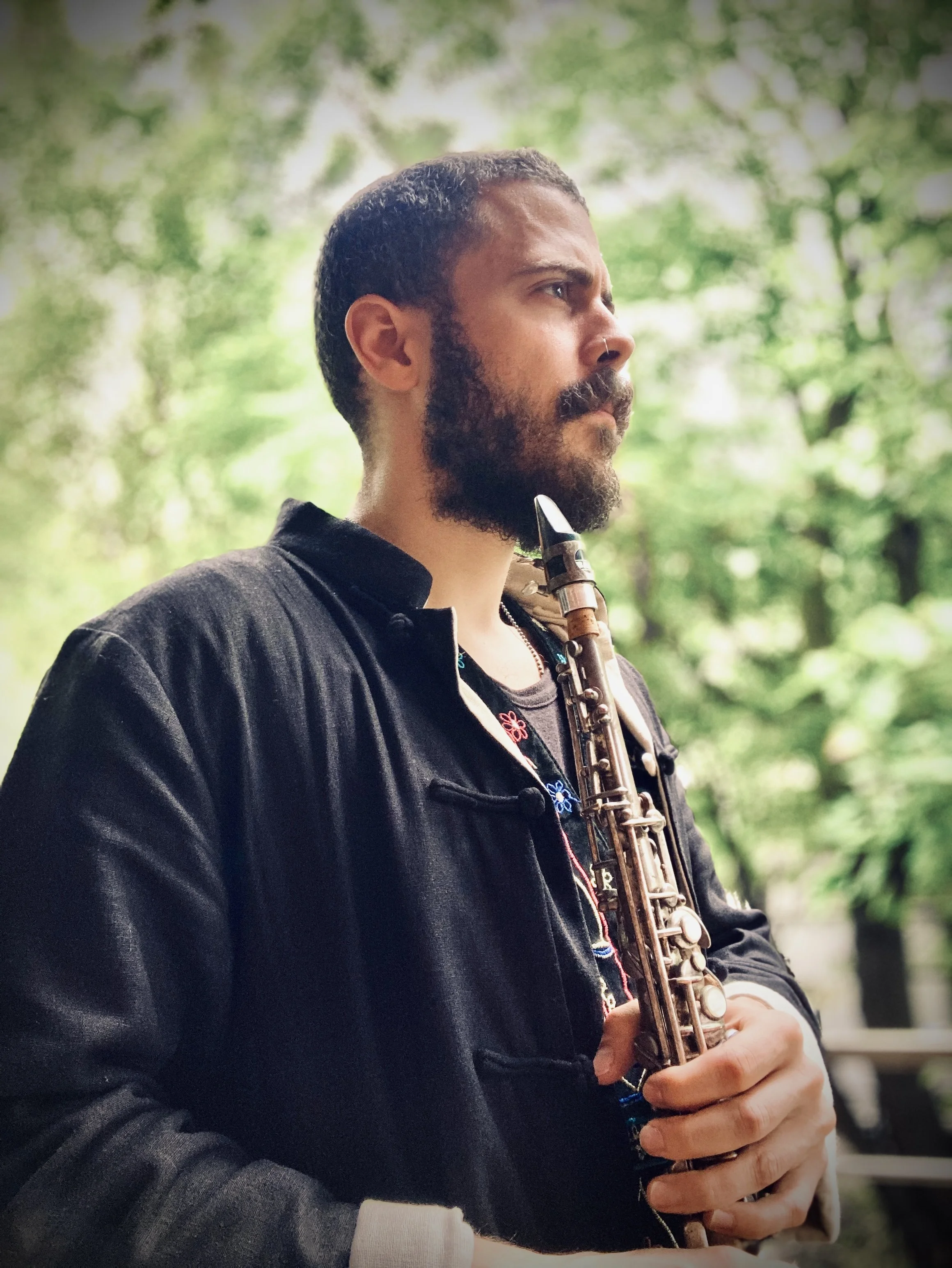 A man with a beard and mustache holding a saxophone, standing outdoors in front of green trees, looking thoughtfully to the side.
