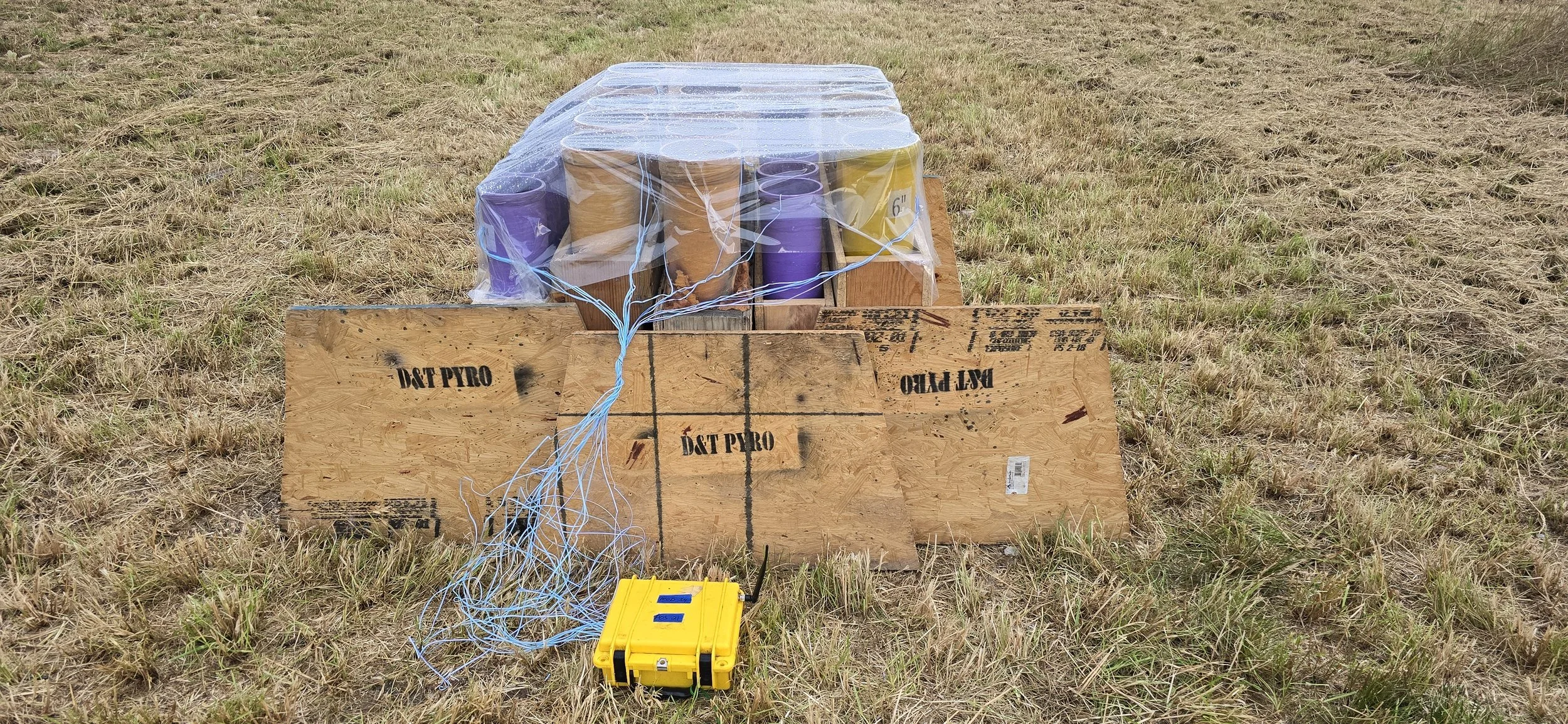 Explosive fireworks on the ground with a yellow safety box and wires, set up outdoors on grass. 1.3 fireworks ready to be lit before a show. Even chance of rain doesn't slow us down!