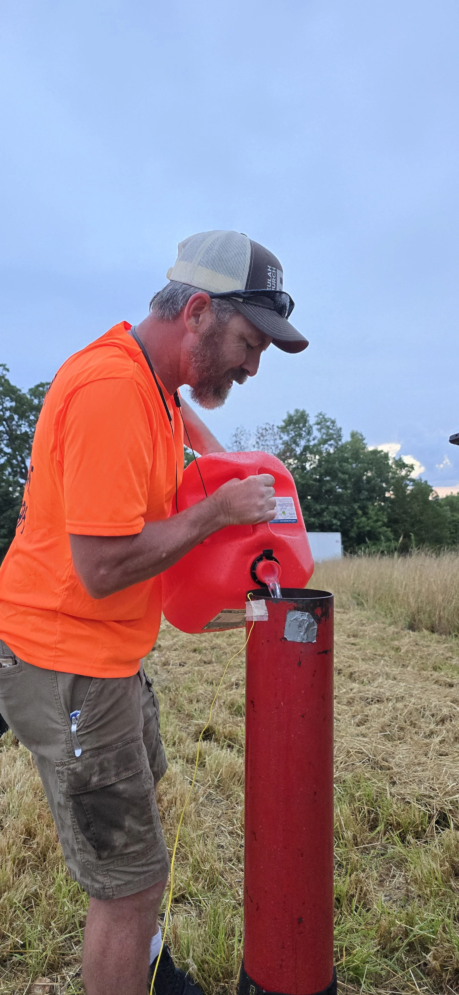 A man with a beard wearing an orange shirt, gray shorts, and a gray and black cap, is filling a red fire extinguisher container with water in an open field during twilight, with trees and a cloudy sky in the background. Josh filling up fireball tubes