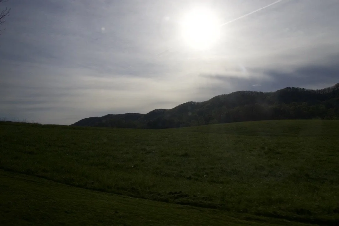 Landscape of green rolling hills with a dark mountain range in the background under a partly cloudy sky illuminated by the sun.