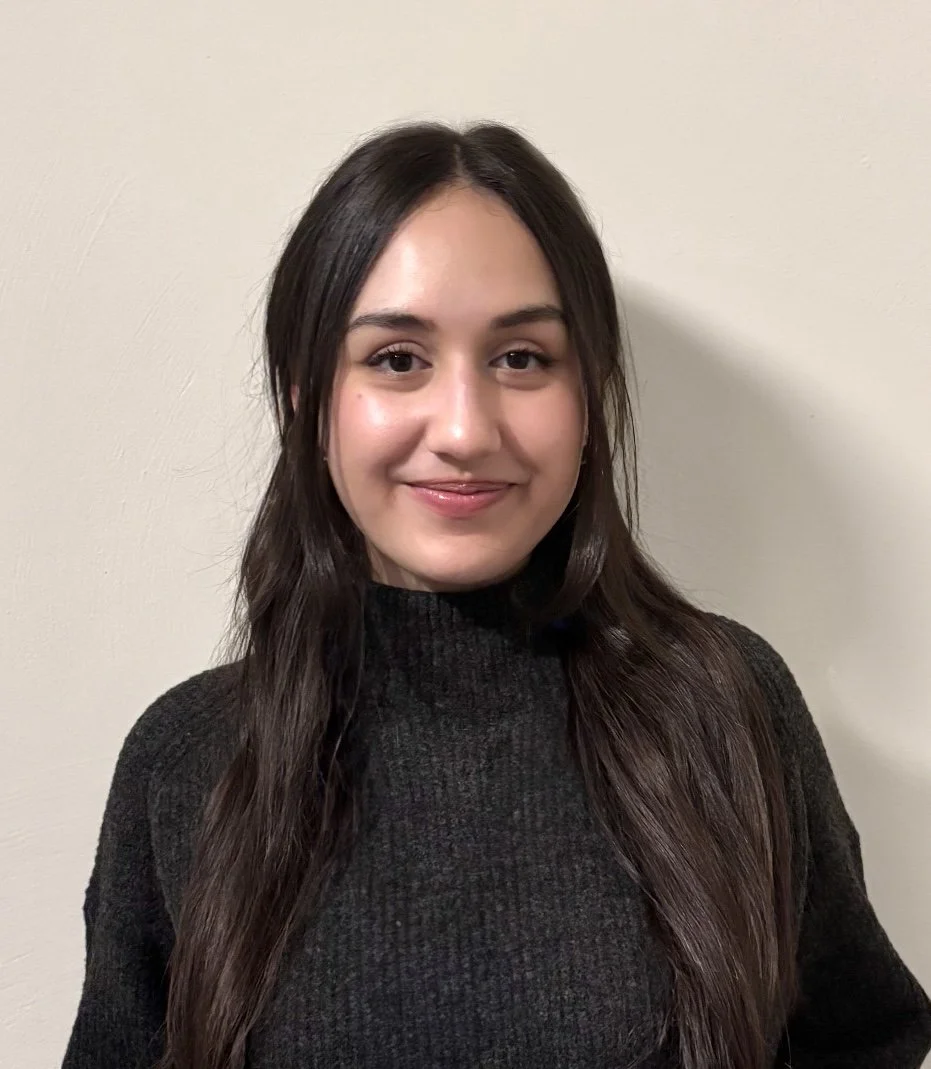 A young woman with long dark brown hair, wearing a black turtleneck sweater, standing in front of a plain cream-colored wall, smiling at the camera.