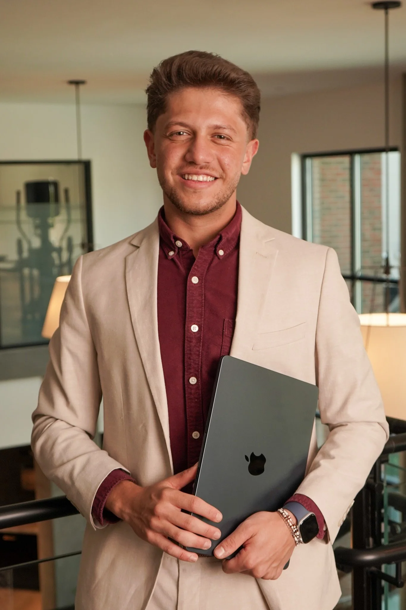 A man in a beige suit and maroon shirt holding a closed laptop with a black Apple logo, standing in a modern office space with large windows and ambient lighting.