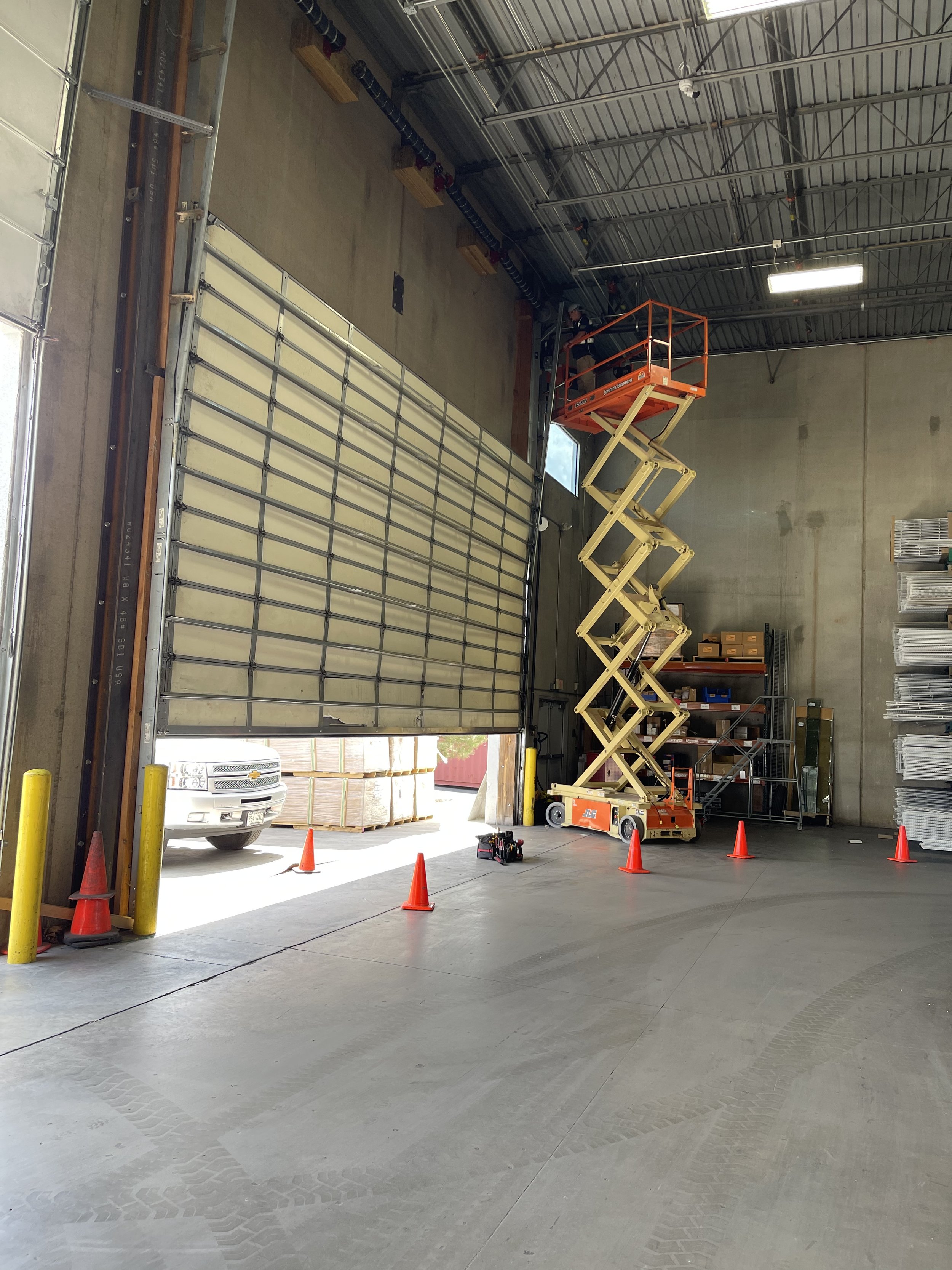 Inside a warehouse with a large open garage door, a scissor lift with a worker reaching near the ceiling, orange traffic cones set around the lift, and organized shelves with boxes and metal racks along the walls.