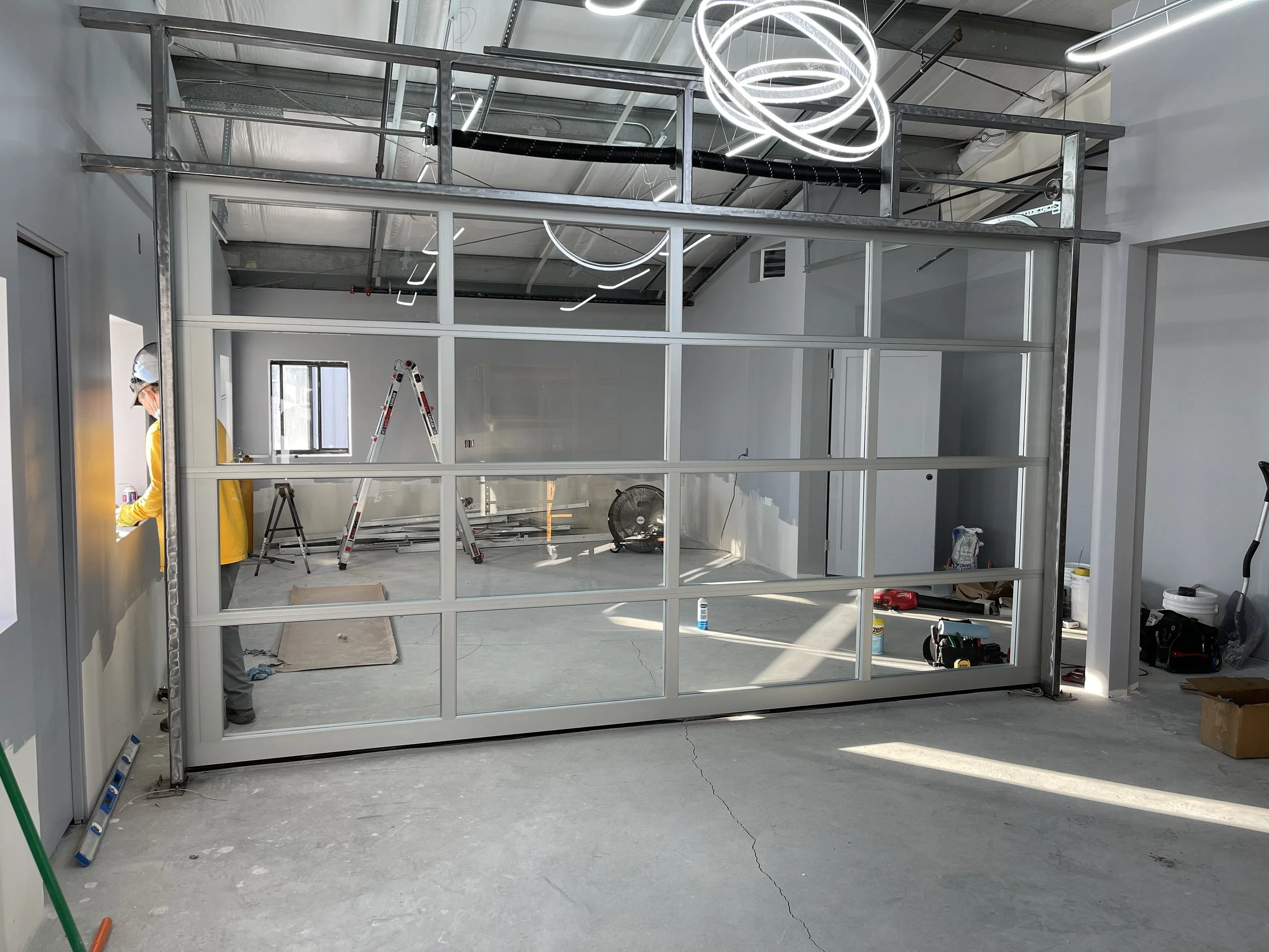 Construction site with a glass garage door installed by Galaxy Doors garage services, a worker in yellow jacket, ladder, and various tools and materials scattered around.
