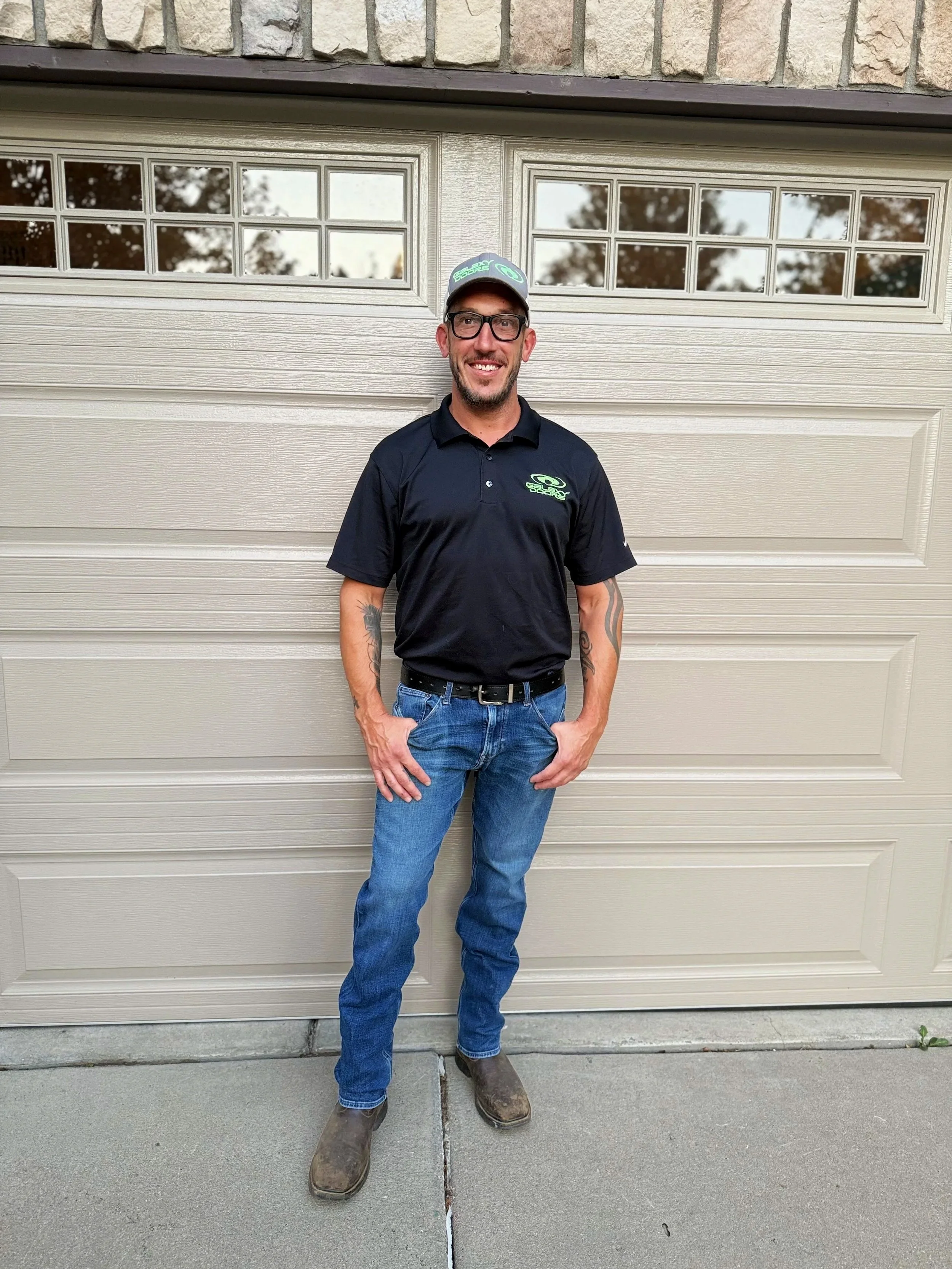 The owner of Galaxy Doors garage repair services, Chris Dimellio, standing in front of a beige garage door, wearing glasses, a black polo shirt with a Galaxy doors logo, jeans, and brown boots.