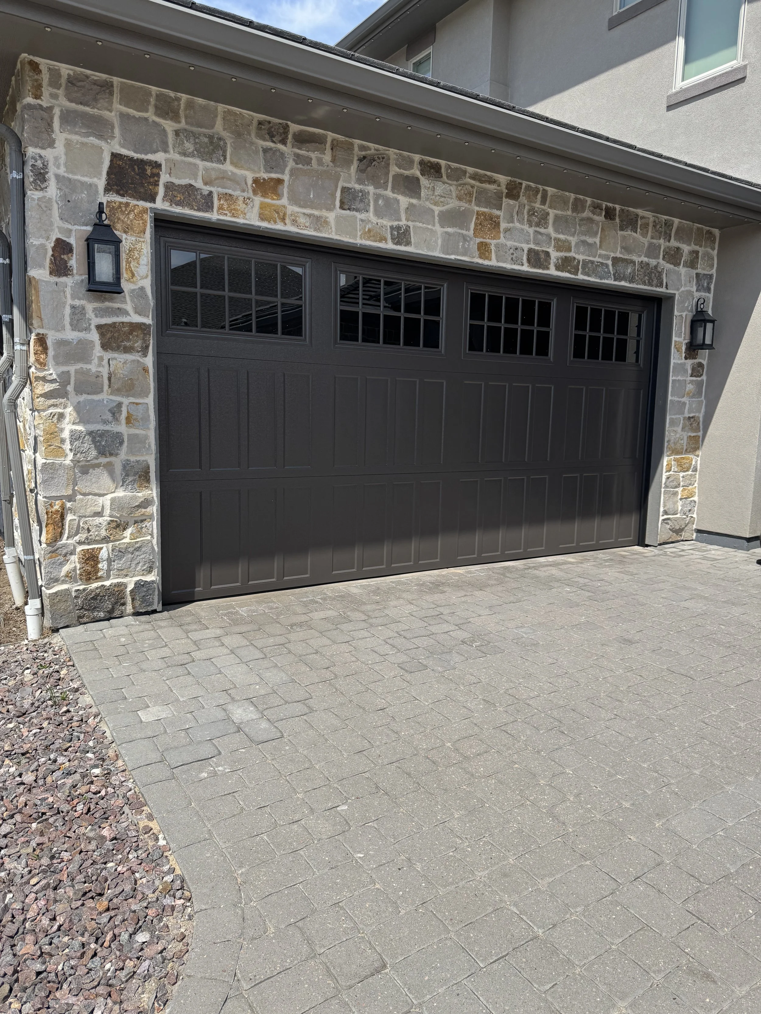 Black garage that was installed by Galaxy Doors garage repair and installation services, with window panes at the top, stone wall on either side, outdoor wall-mounted lanterns, paved driveway, and part of a house with gray siding.