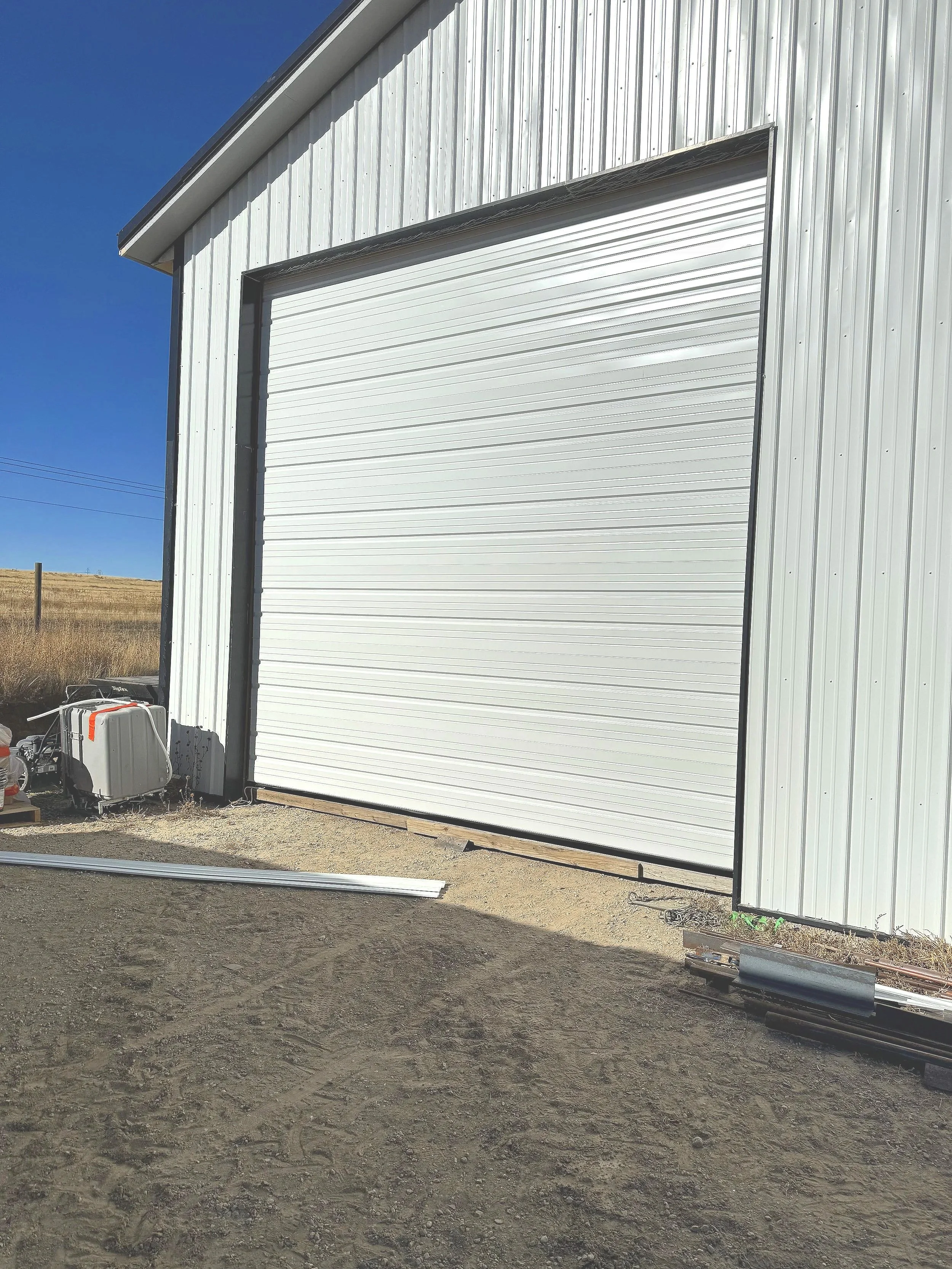 White metal building with a large roll-up garage door, installed by Galaxy Doors garage repair services, outdoors on a sunny day. There are construction materials and equipment in front of the building on a dirt ground.