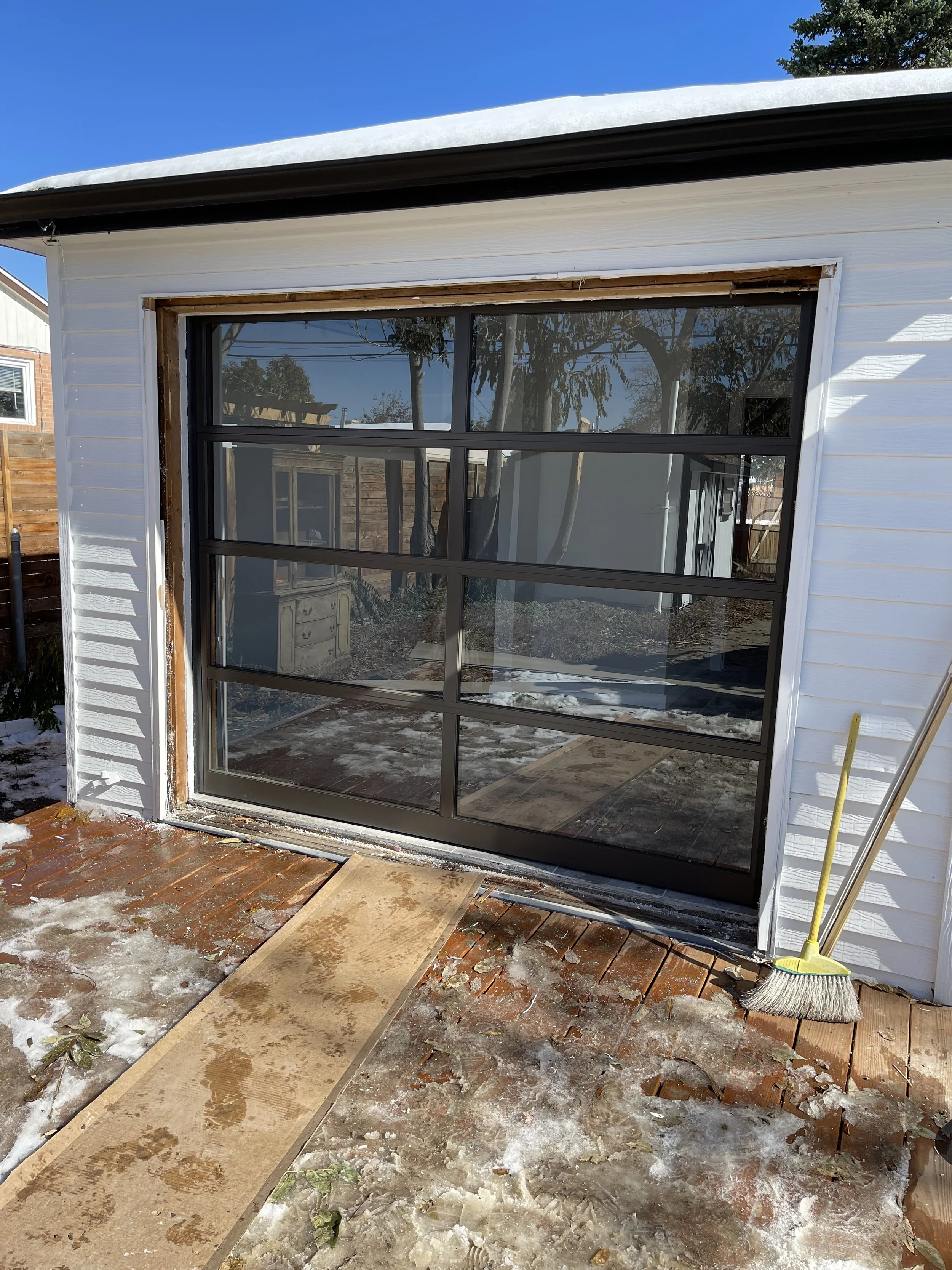 A large black-framed glass garage door, partially under construction or repair, with a wooden walkway and snow on the ground. A broom leans against the wall on the right.