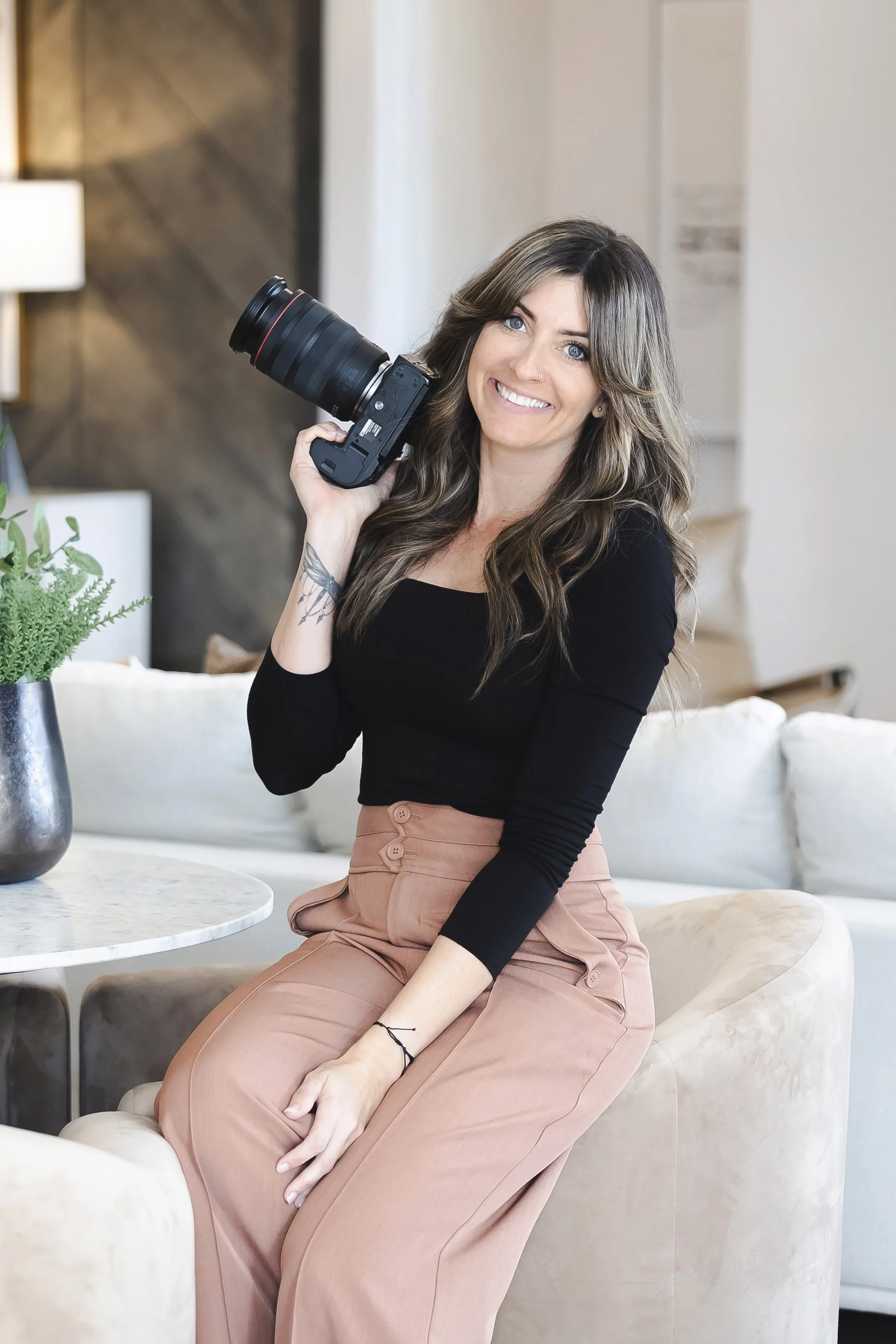 Jennifer Lueck with long brown hair and blue eyes, smiling, leaning on a kitchen island with a camera hanging from her arm, in a modern kitchen with stone and wood decor.