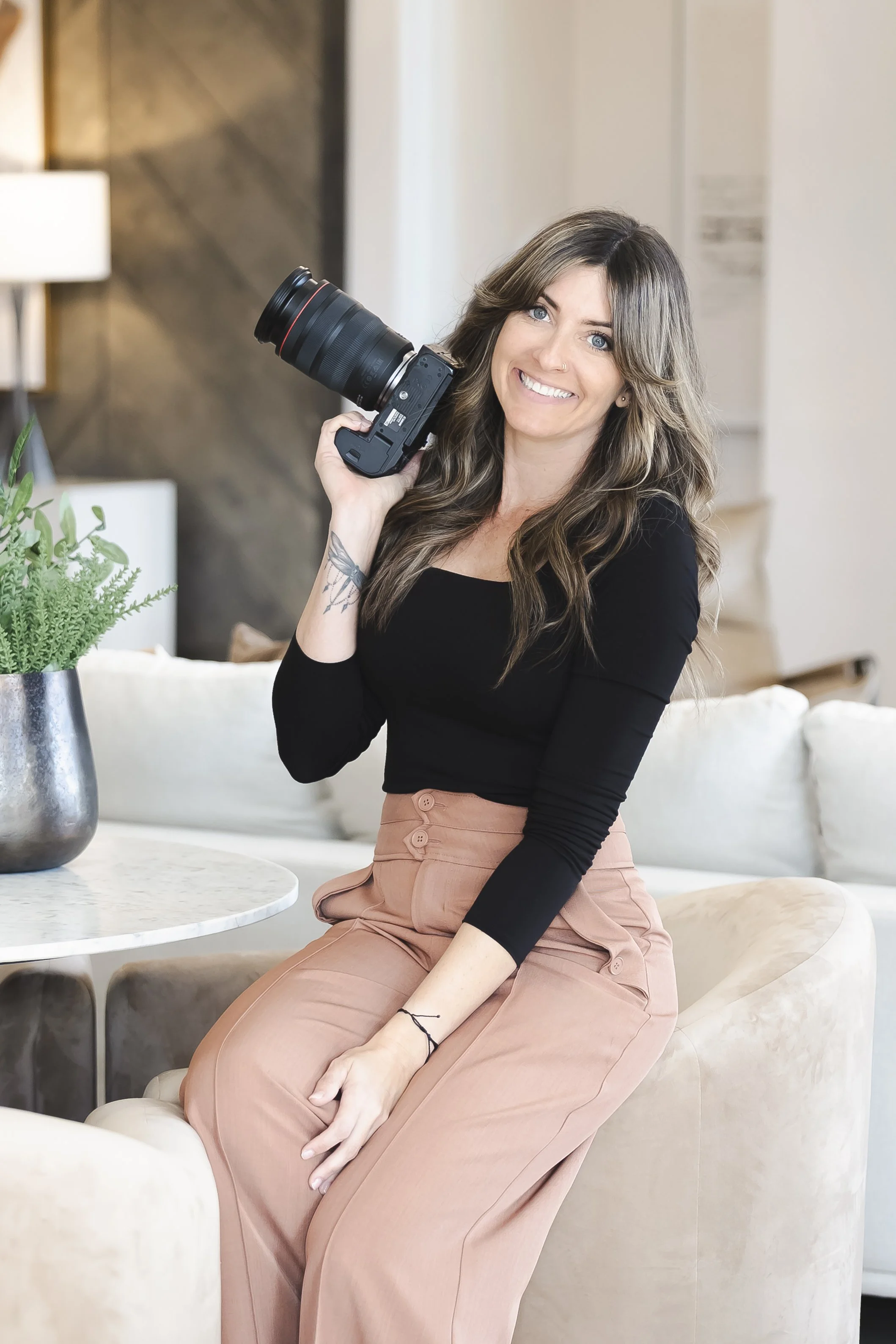Jennifer Lueck with long brown hair and blue eyes, smiling, leaning on a kitchen island with a camera hanging from her arm, in a modern kitchen with stone and wood decor.